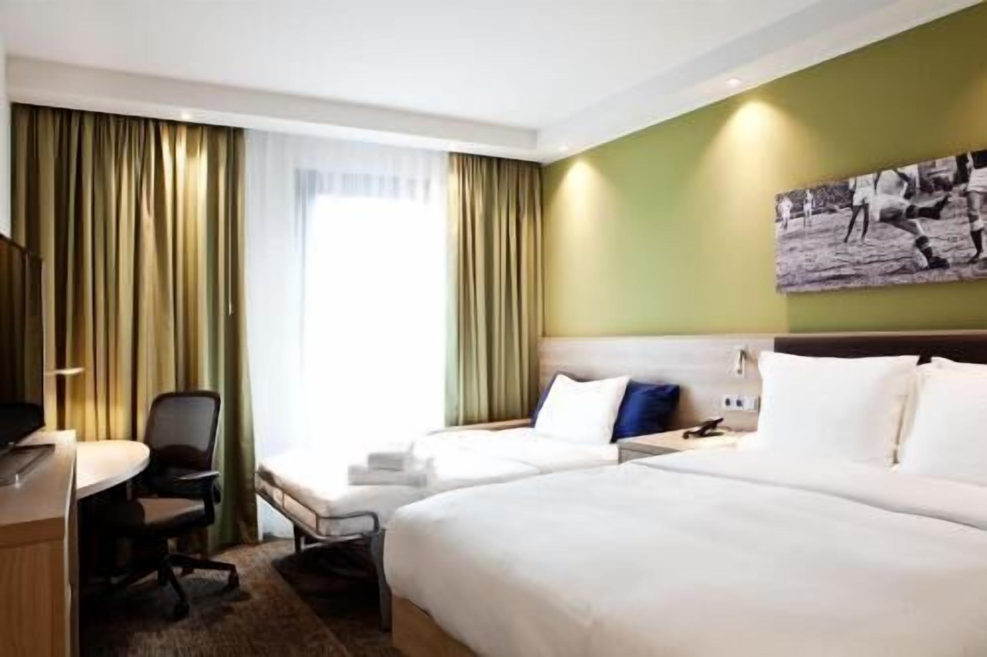 A hotel room with two beds, a desk, a chair, and a large window with curtains.
