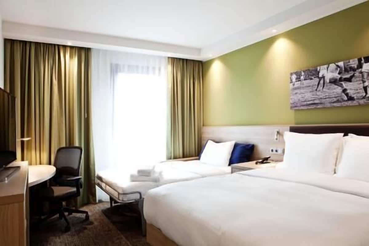Ein Hotelzimmer mit zwei Betten, einem Schreibtisch, einem Sessel und einem großen Fenster mit Vorhängen.