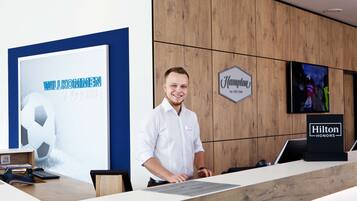 A hotel reception desk with a "Hilton Honors" sign, a welcome sign, and a television screen displaying a sports event.