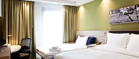 Quarto de hotel com duas camas, uma escrivaninha com cadeira, televisão e uma janela grande com cortinas.