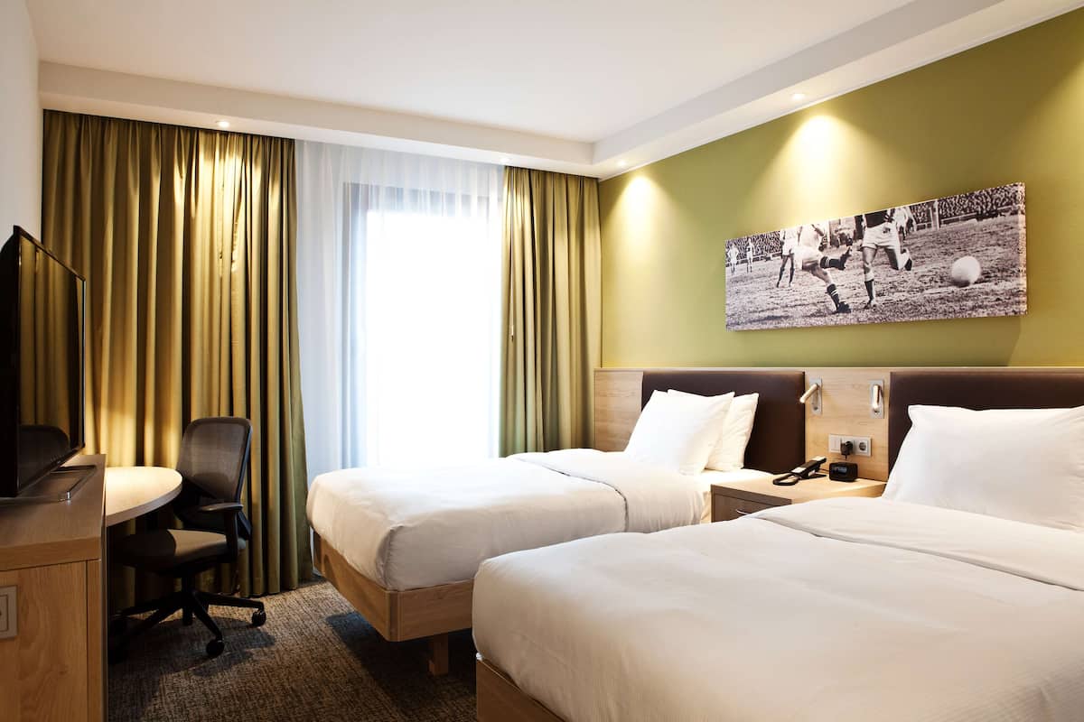 Ein Hotelzimmer mit zwei Betten, einem Schreibtisch, einem Sessel, einem Fernseher und einem Wandbild.