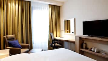 A hotel room with a bed, a desk with a flat-screen TV, a chair, and a large window with curtains.
