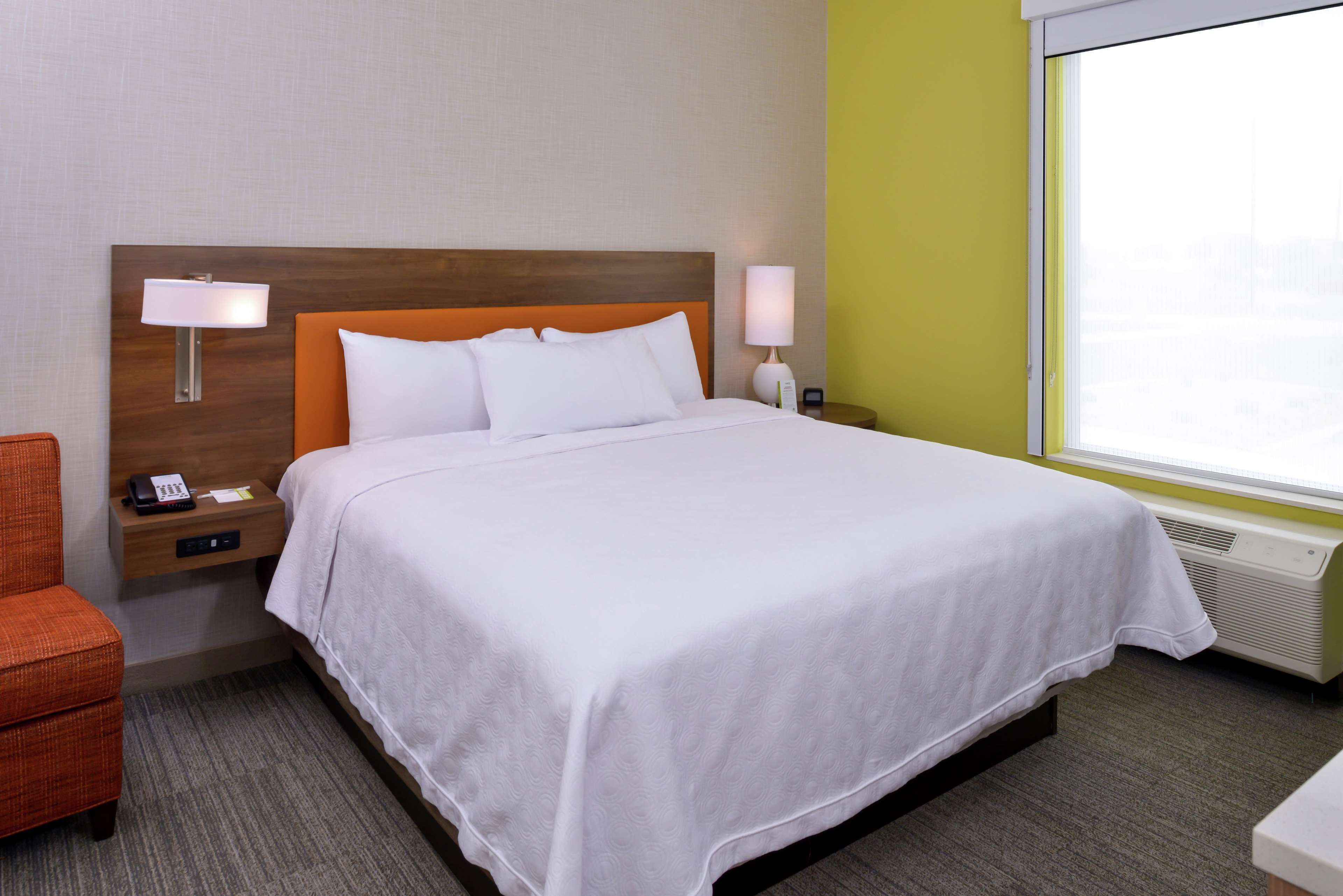 A hotel room with a bed, a nightstand with a lamp, a chair, and a window with a view.