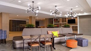 A modern living room with a sectional sofa, two wooden chairs, a coffee table, and a TV mounted on the wall. - Home2 Suites by Hilton Merrillville (Merrillville)