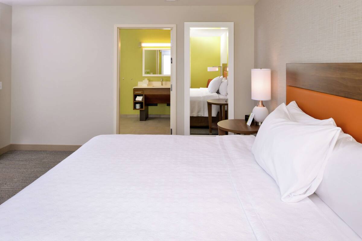 Hotelkamer met een groot bed, een nachtkastje met lamp, een badkamer zichtbaar door een geopende deur en een spiegel boven de wastafel.