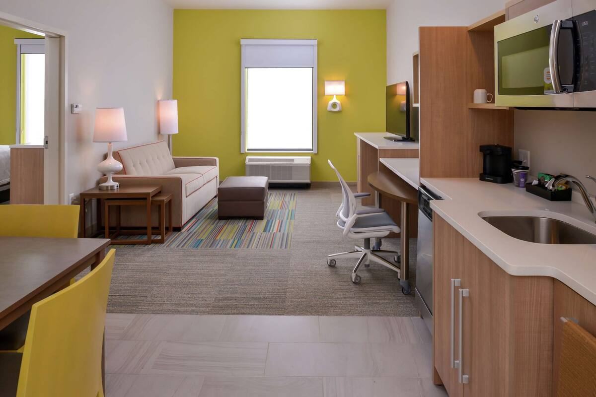 Een moderne hotelkamer met een kitchenette, een bank, een eettafel en een raam met zonwering.