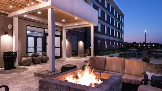 An outdoor seating area with a fire pit, a covered patio, and a building in the background. - Home2 Suites by Hilton Merrillville (Merrillville)