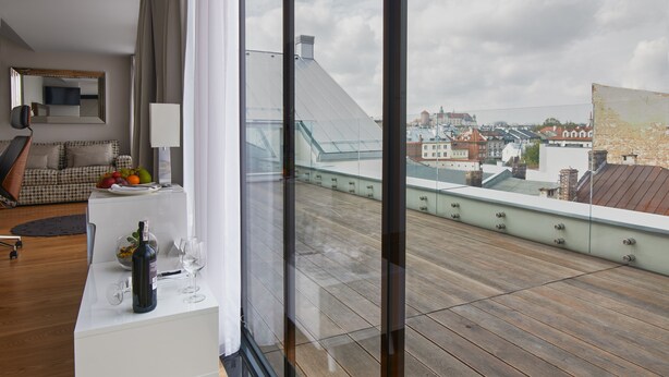 A modern hotel room with a wooden floor, a white console table with a bottle and glasses, a sofa, and a view of the cityscape through large windows. - INX Design Hotel (Kraków)