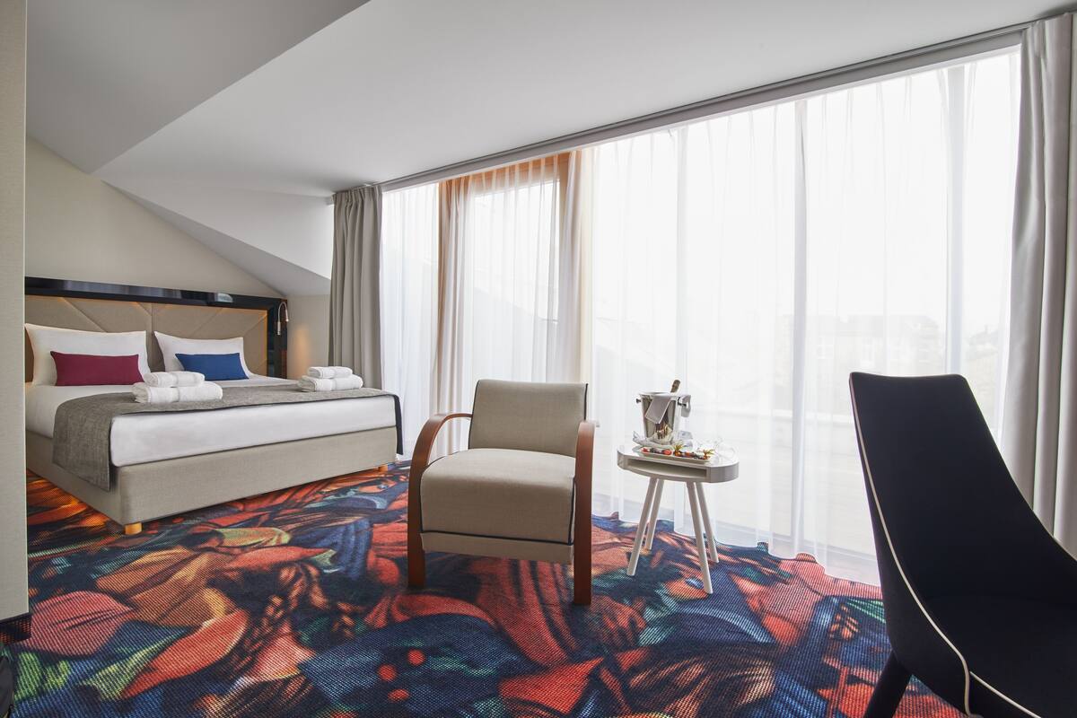 Una habitación de hotel con una cama, una silla, una mesita pequeña y ventanas grandes con cortinas.