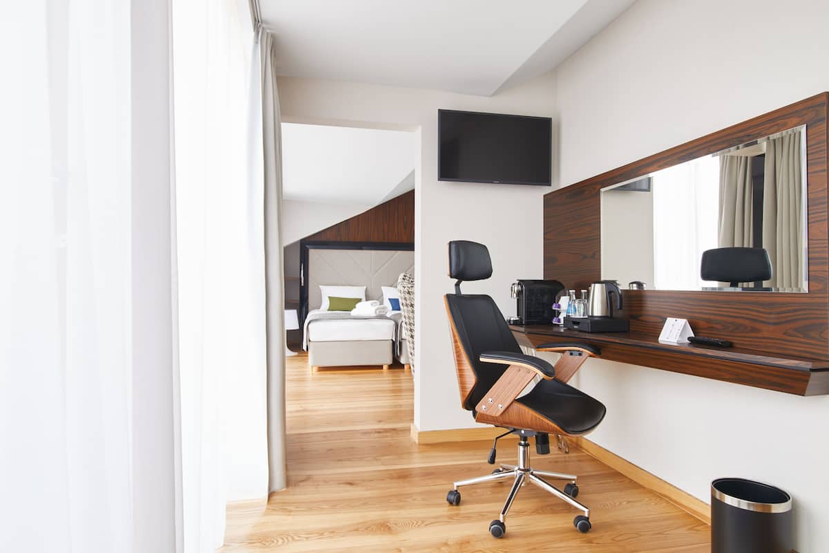Una habitación de hotel moderna con suelo de madera, un escritorio con computadora, una silla, una cama con cabecera blanca y un televisor montado en la pared.