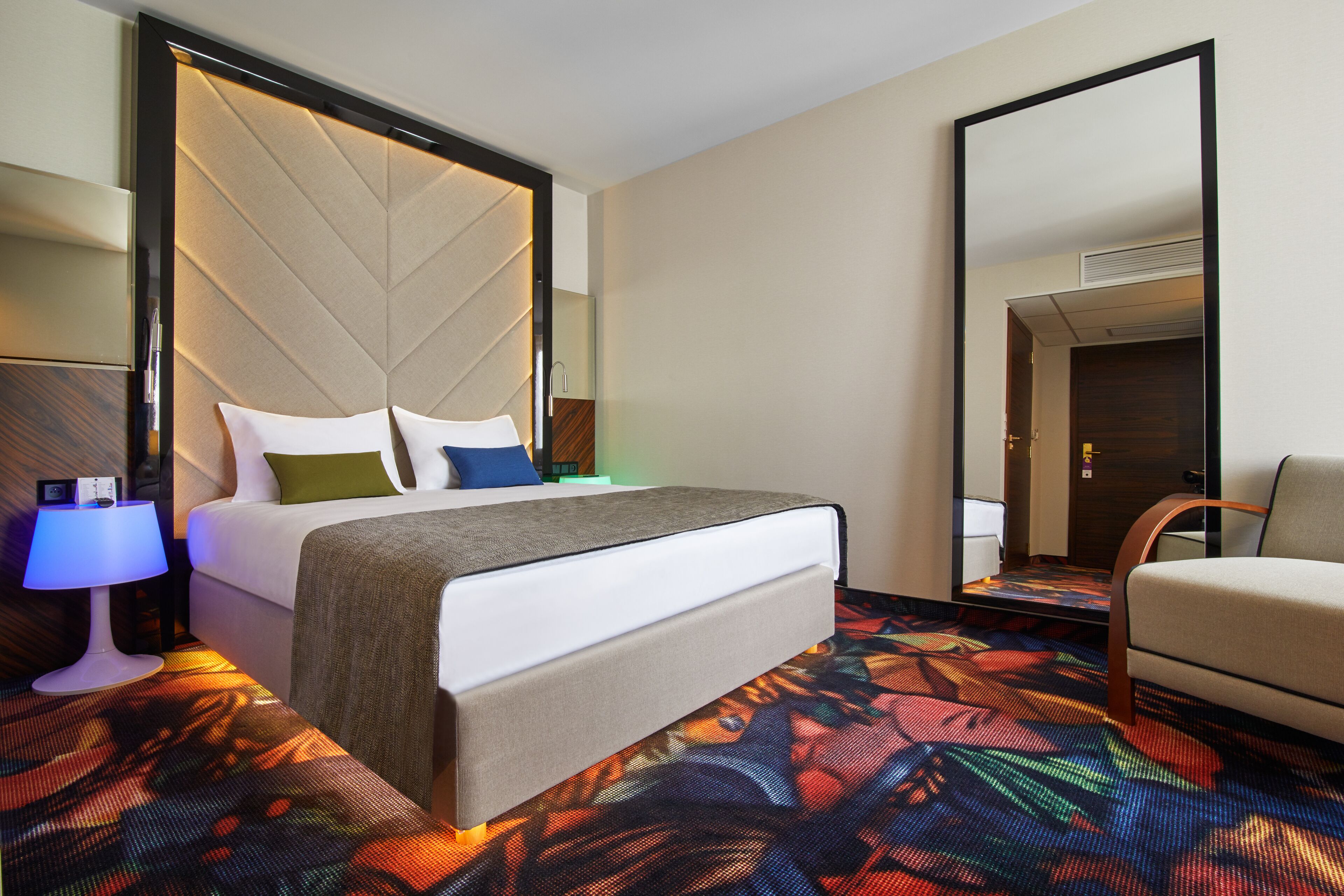 Una habitación de hotel con una cama, una silla, una mesita de noche, una lámpara y un espejo.