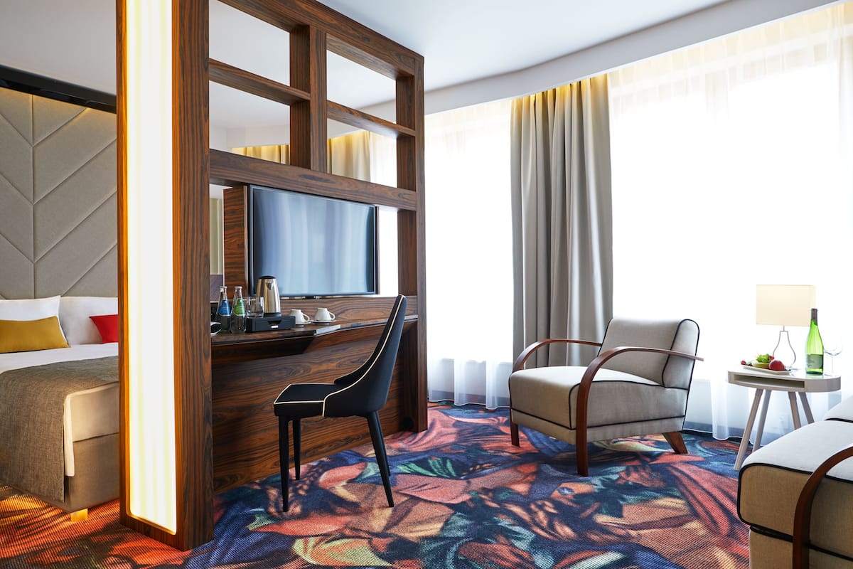 Habitación de hotel con cama, escritorio con silla, televisor, mesita de noche con lámpara y ventana con cortinas.