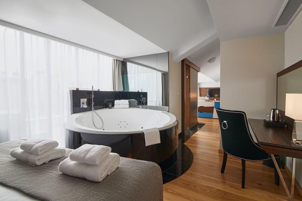 Una habitación de hotel moderna con una bañera grande, un comedor con mesa y sillas, y vista a la cocina en el fondo.