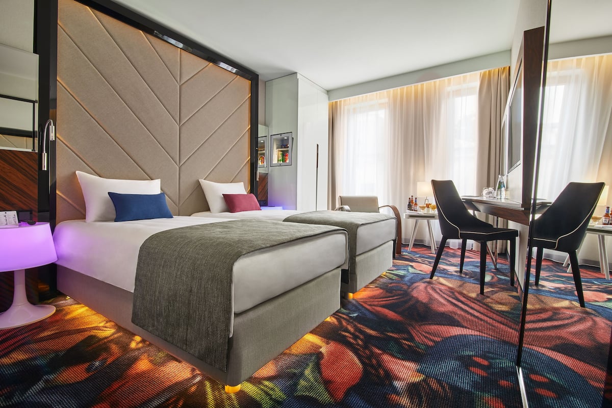 Habitación de hotel con cama, dos mesitas de noche, un escritorio con sillas, un espejo grande y una alfombra colorida.