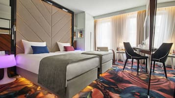 Habitación de hotel con cama, dos mesitas de noche, un escritorio con sillas, un espejo grande y una alfombra colorida.