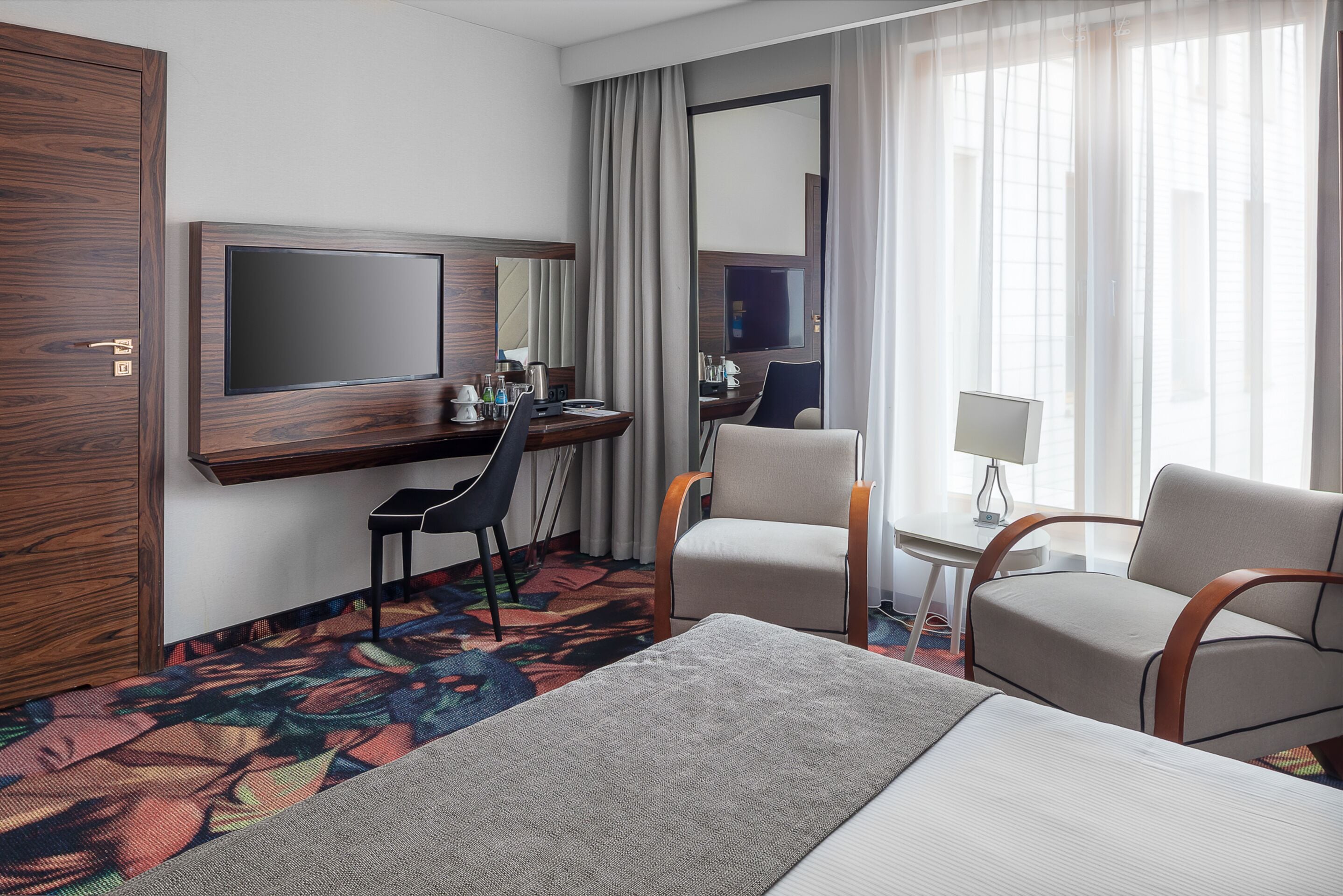 A hotel room with a bed, two armchairs, a desk with a chair, a television, and a large mirror.