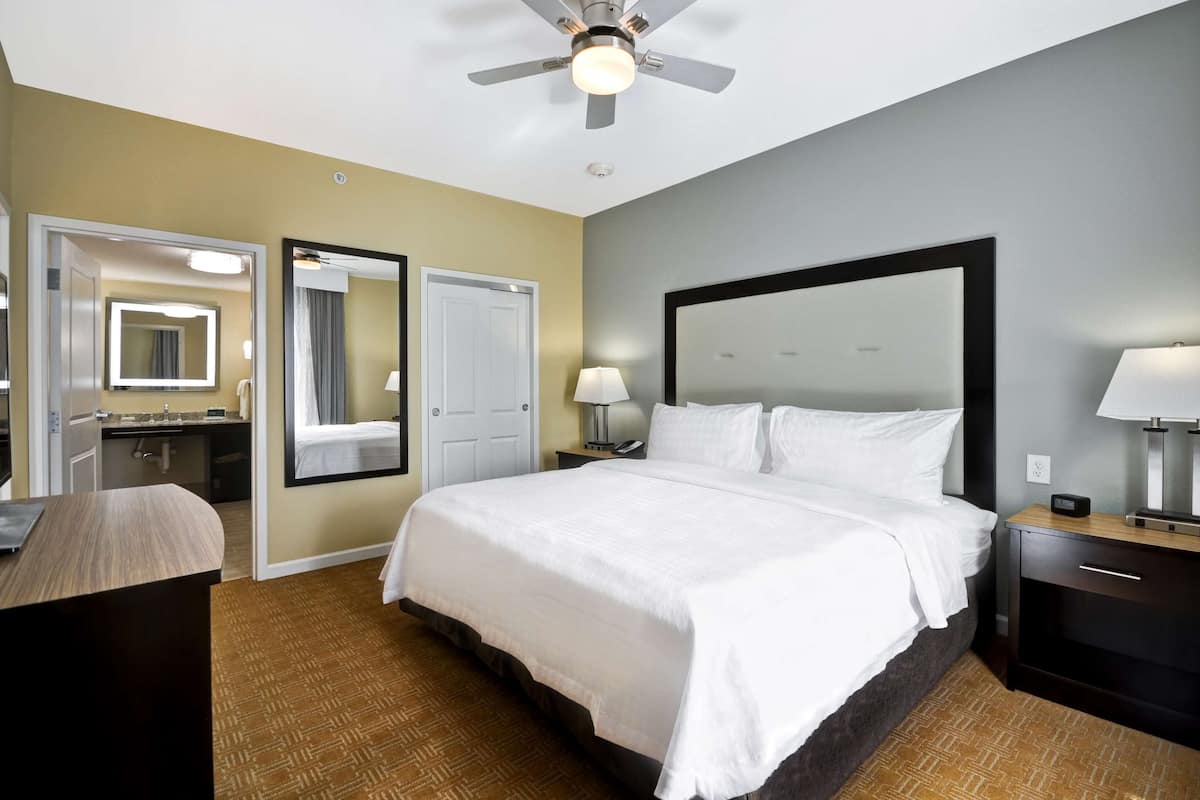 A hotel room with a large bed, a desk, a chair, a ceiling fan, and a bathroom visible through an open door.