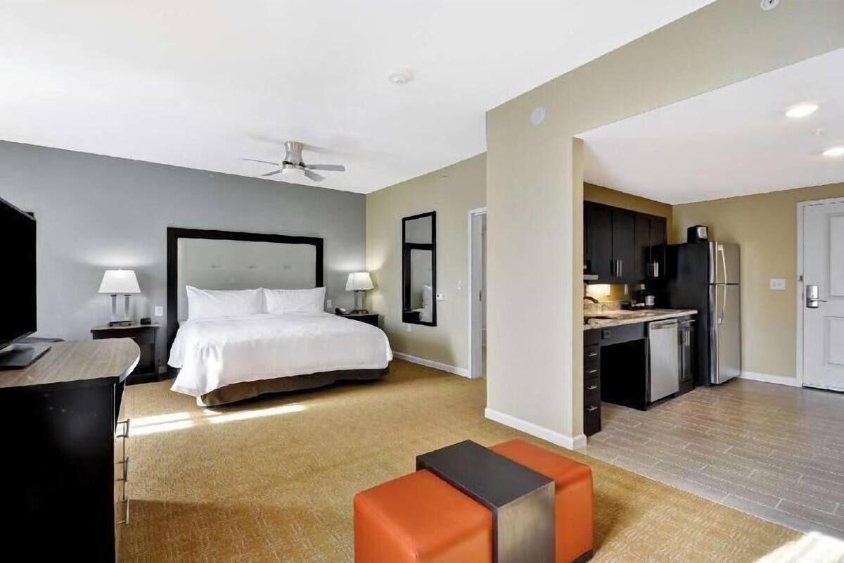 A hotel room with a bed, a desk, a chair, a TV, a ceiling fan, a kitchen area, and a bathroom.