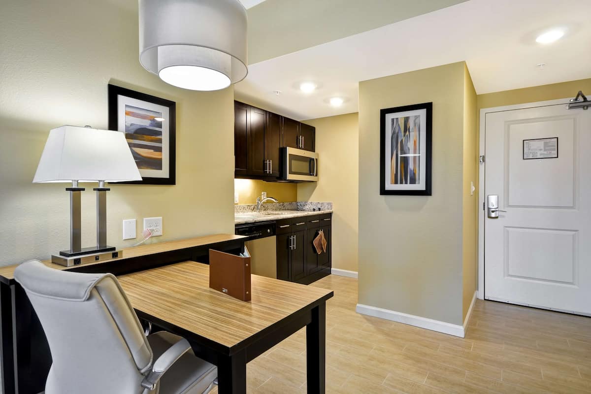 A modern kitchen with dark cabinets, a microwave, and a sink. There is a dining table with a chair and a lamp. The walls are painted in a light color, and there is a door visible.