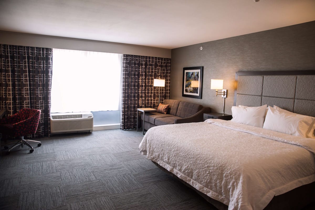 Habitación de hotel con una cama grande, un sofá, un escritorio y una silla. Se ve una ventana con cortinas y un equipo de aire acondicionado.