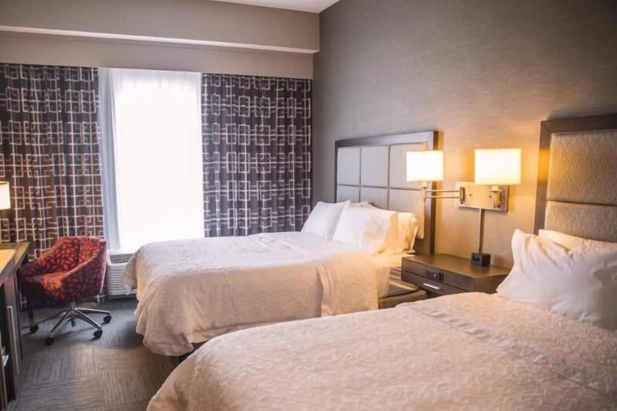 Una habitación de hotel con dos camas, un escritorio, una silla y un ventanal con cortinas.