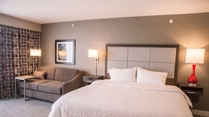 A hotel room with a large bed, a sofa, a nightstand, and a lamp. - Hampton Inn Pontiac (Pontiac)