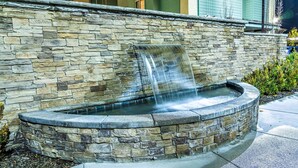 A stone water feature with a waterfall, surrounded by a stone wall and a paved area.