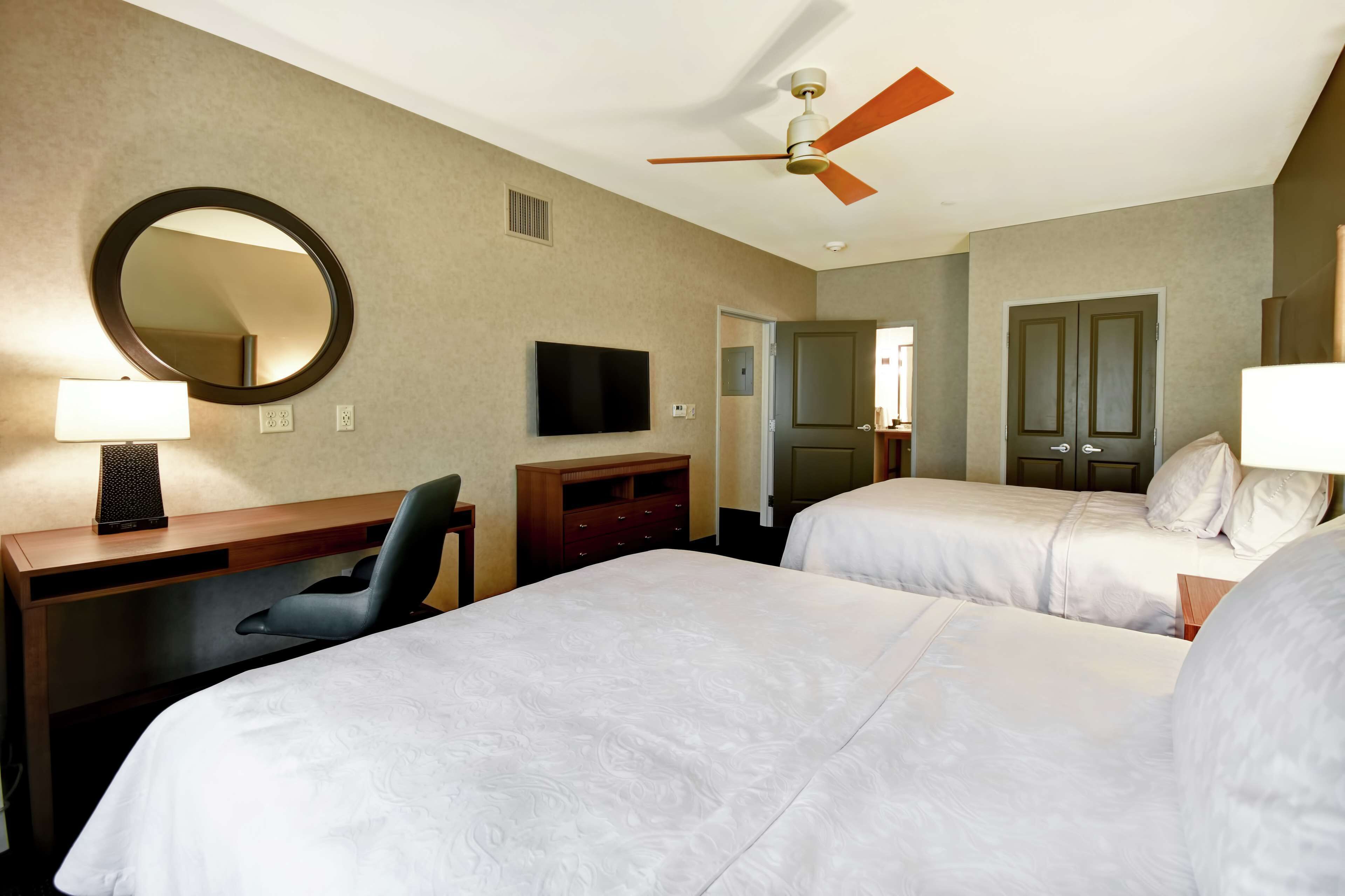 Une chambre d’hôtel avec deux lits, un bureau, une chaise, une télévision, un ventilateur de plafond et un miroir.