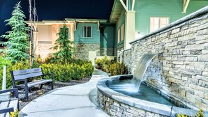 A stone water feature with a waterfall, a concrete pathway, and benches in a landscaped area with evergreen trees and shrubs.