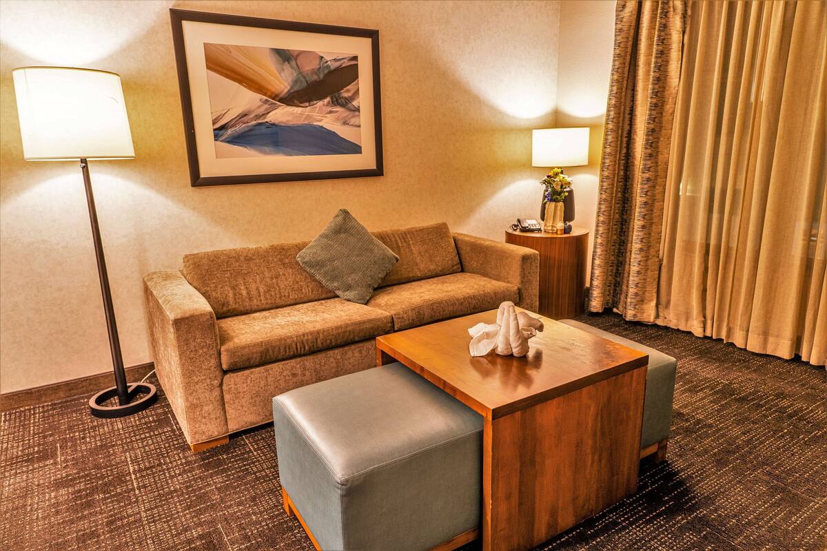 Een hotelkamer met een bank, een houten bijzettafeltje, een nachtkastje met een lamp, een lijst met een afbeelding aan de muur, en een raam met gordijnen.