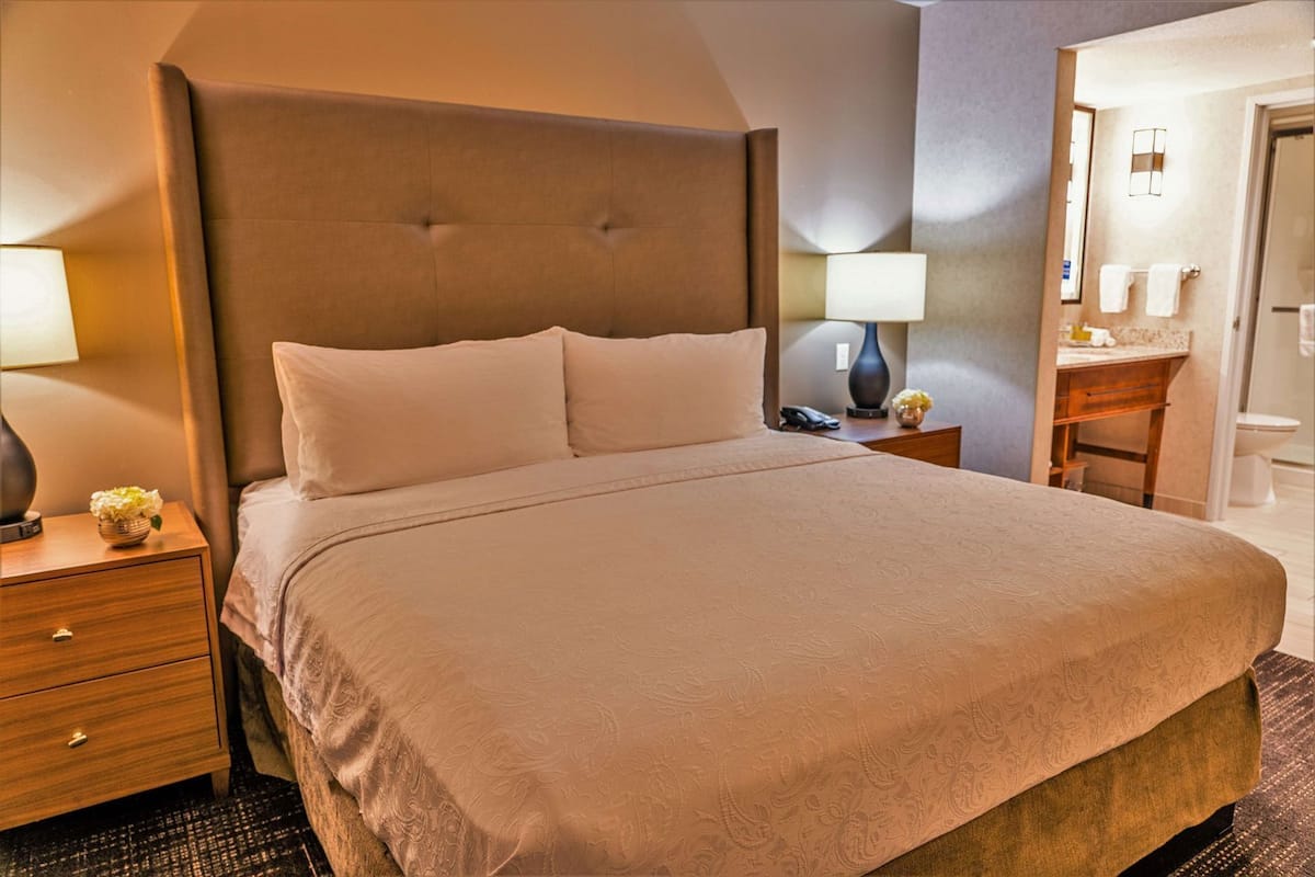 Habitación de hotel con una cama grande, una mesita de noche con lámpara, una mesita auxiliar con un jarrón de flores y un baño visible a través de una puerta abierta.