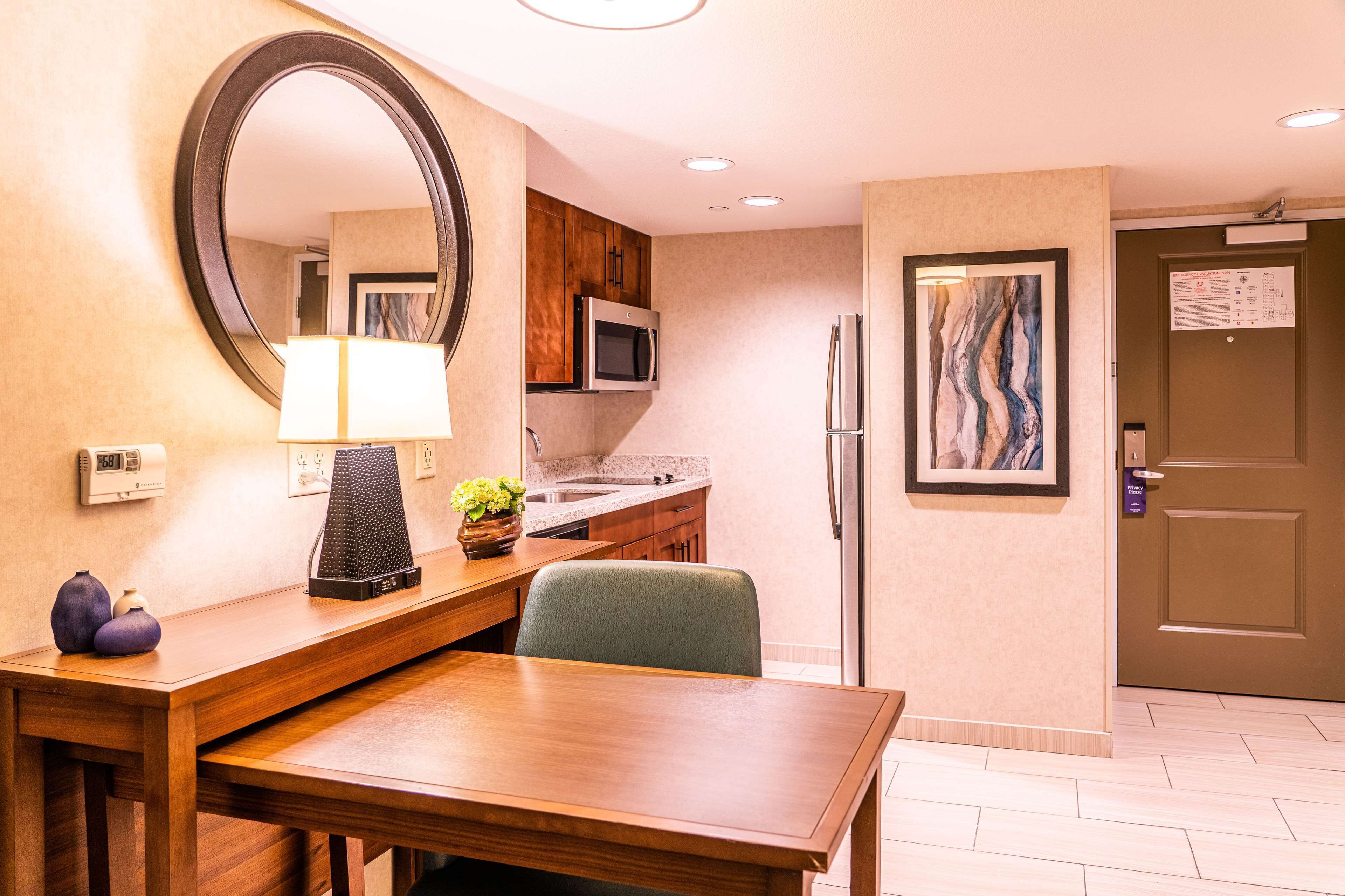 A hotel room with a wooden dining table, a microwave, a refrigerator, a sink, and a large mirror.