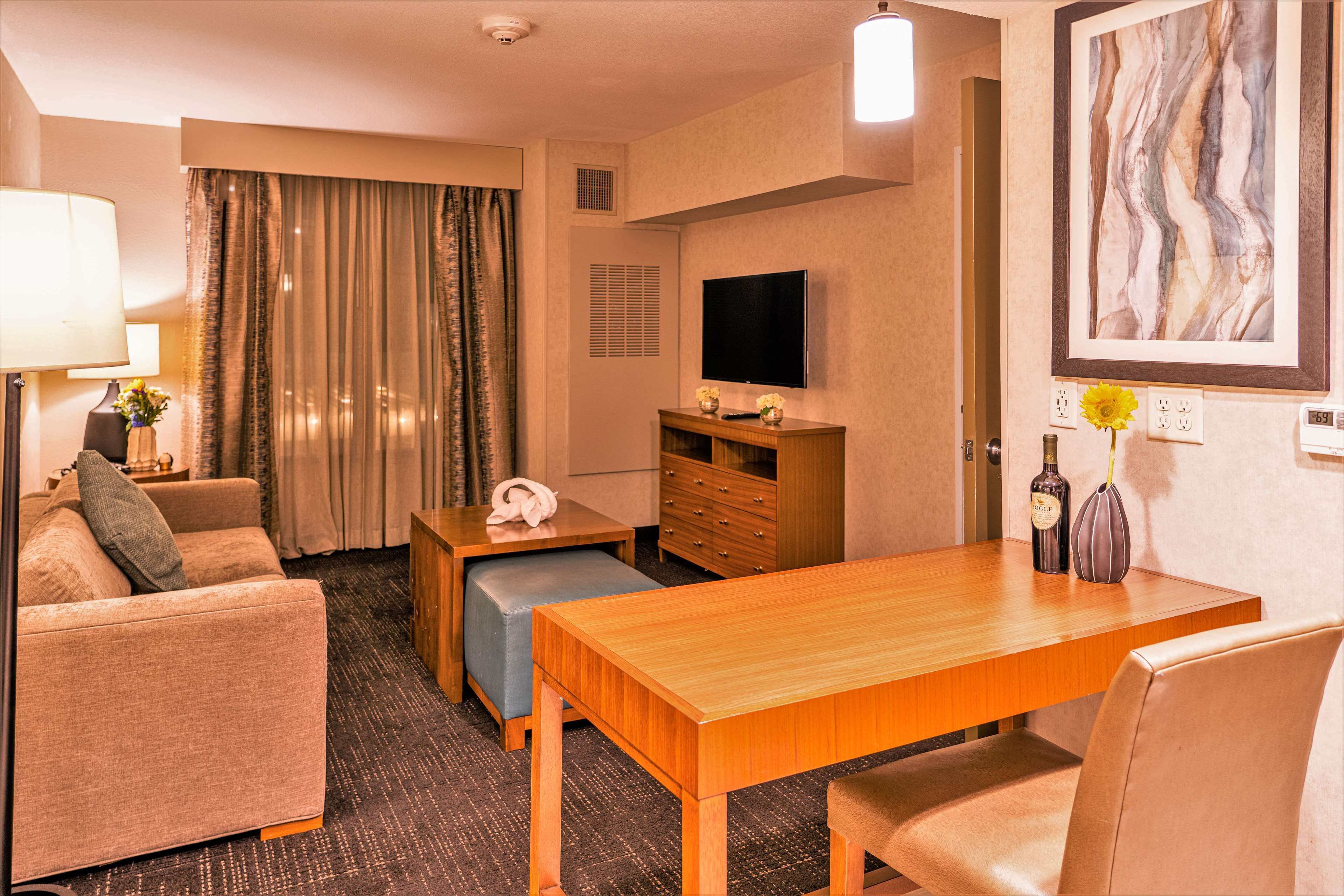 A hotel room with a sofa, a wooden dining table, a TV, a painting, and a lamp.
