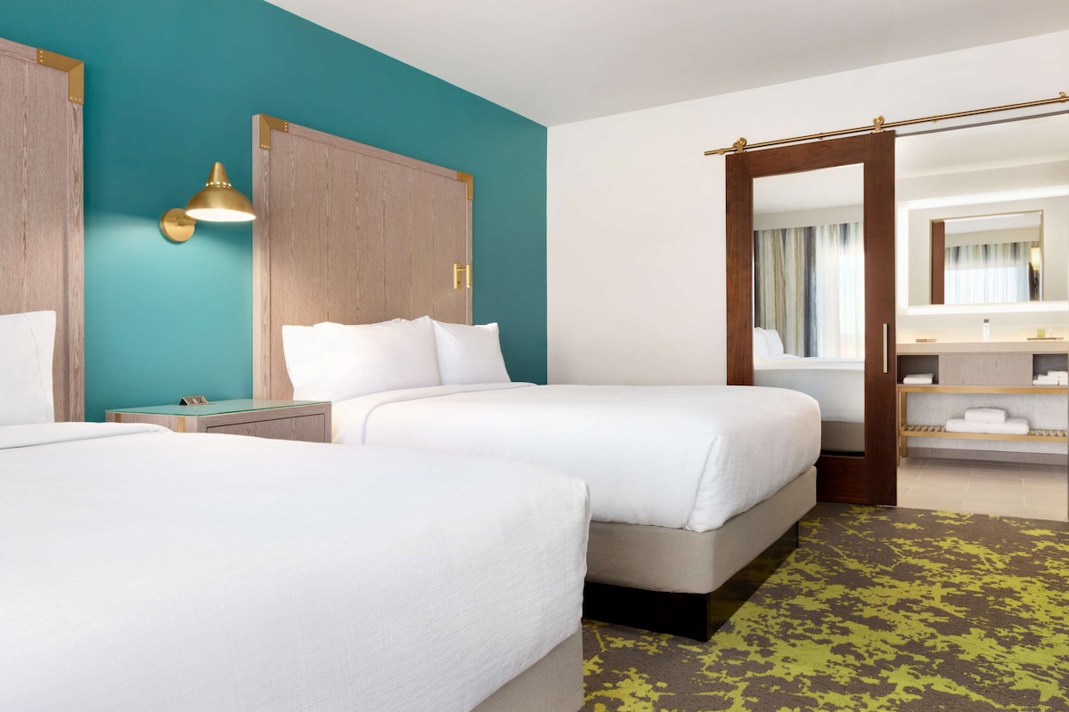 Habitación de hotel con dos camas, un armario de madera, un baño con espejo y un tapete verde estampado.