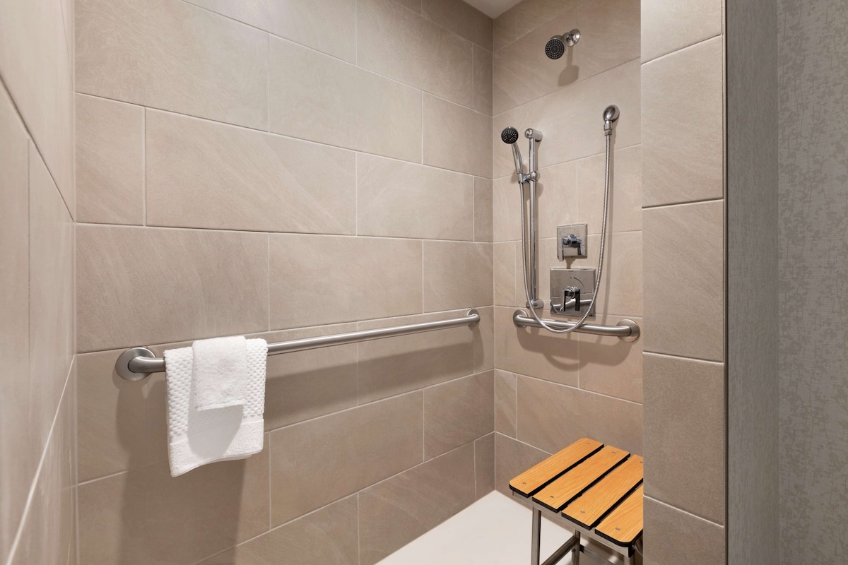 Zona de ducha con azulejos color beige, una barra de apoyo, un perchero, un cabezal de ducha manual y un taburete de madera pequeño.