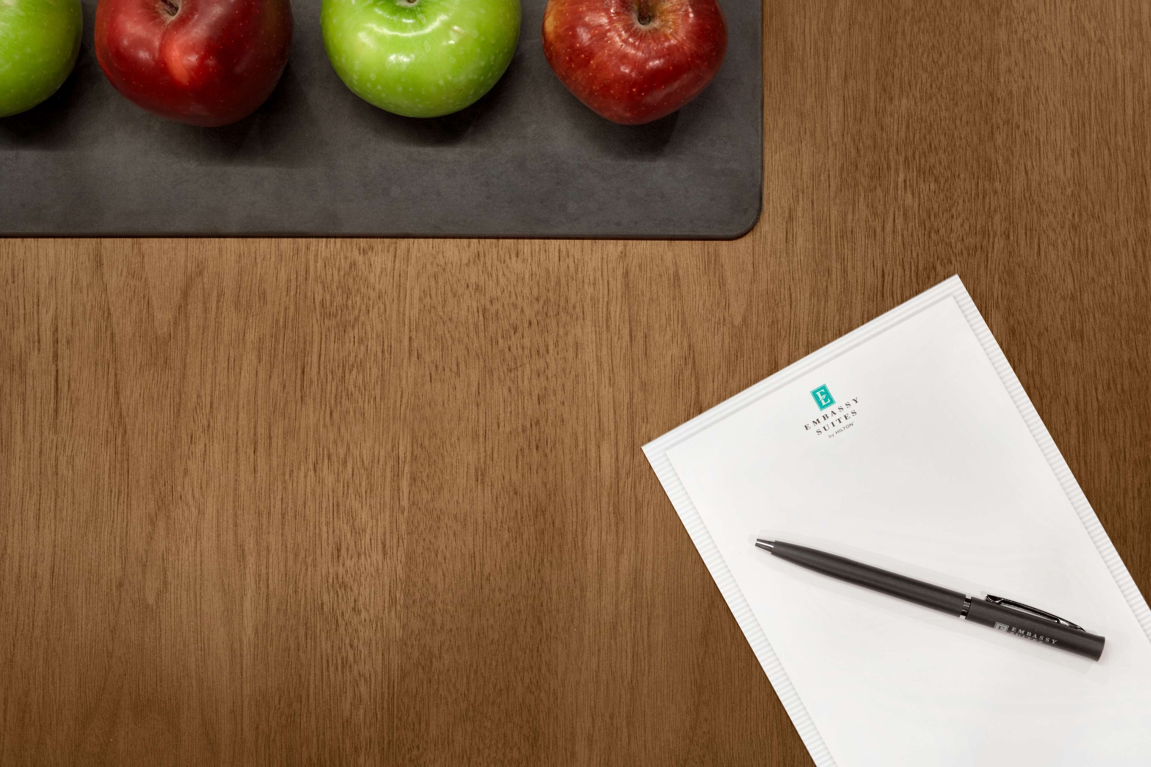 A wooden table with a black mat, three apples, and a pen on a white notepad.