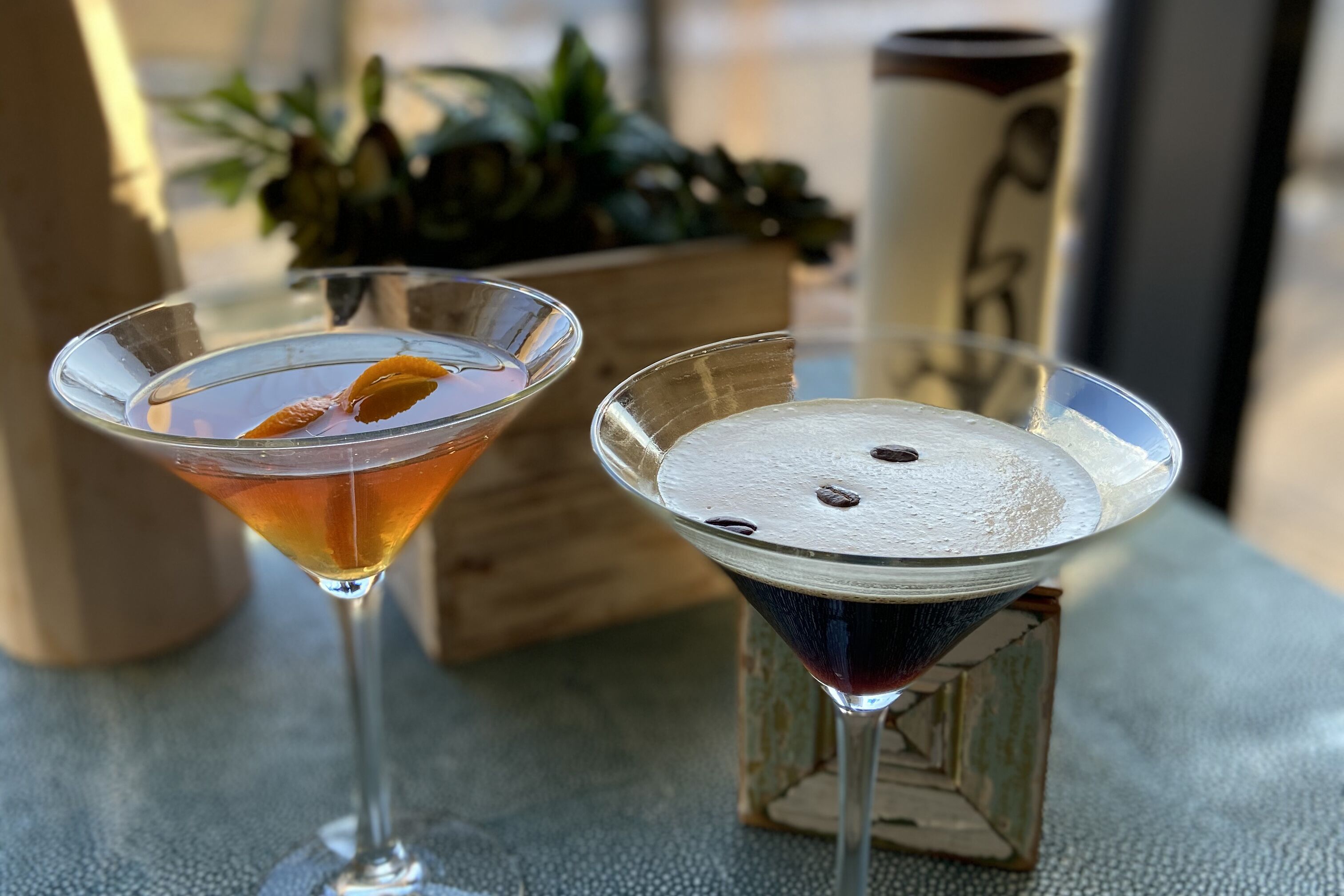 Dos copas de martini sobre una mesa texturizada, con un bloque de madera y una planta de fondo.