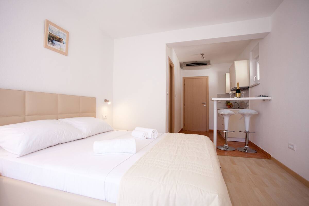 A hotel room with a large bed, a small table with two stools, a mirror above the table, and a door leading to another room.