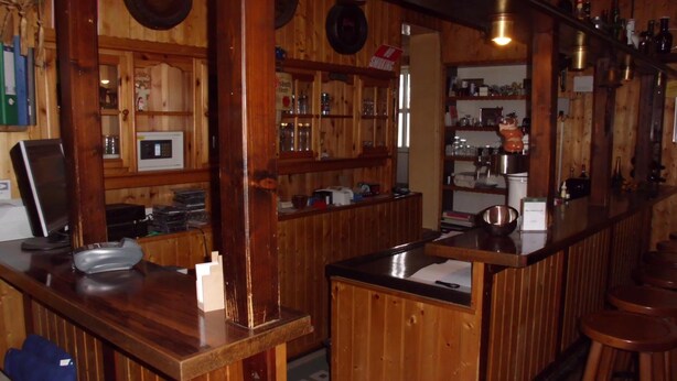 Zona de bar amb barra, tamborets i estanteries plenes de garrafes i vasos.