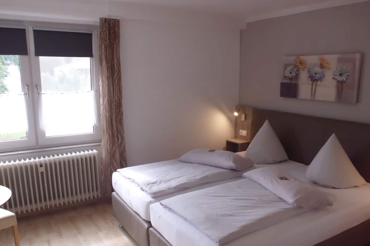 Et hotelværelse med to senge, en radiator, et vindue med gardiner, et bord og et billede hængende på væggen.