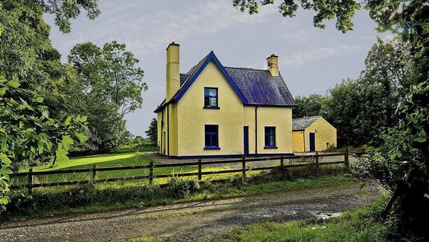 A two-story yellow house with a blue roof and chimney, surrounded by greenery and a wooden fence. - The Gardener's Cottage (Kilvarnet)