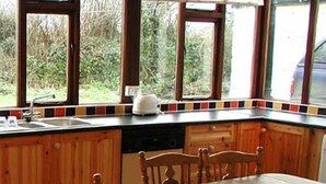A kitchen with wooden cabinets, a dining table, and a view of greenery outside. - The Gardener's Cottage (Kilvarnet)