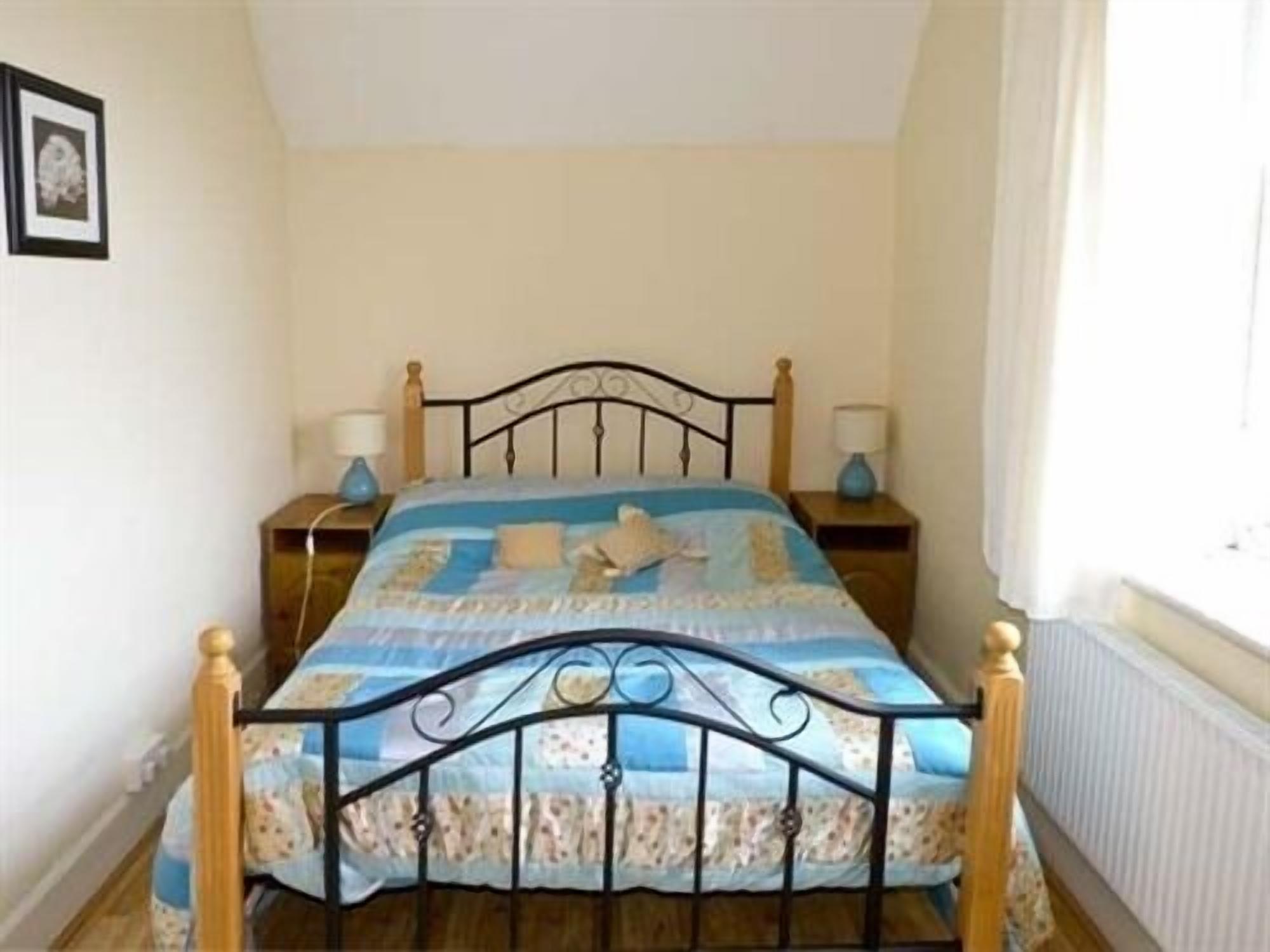 A single bed with a metal frame, a blue and white patterned bedspread, two bedside tables with lamps, and a window with curtains.