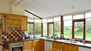 A kitchen with wooden cabinets, a tiled backsplash, and a large window overlooking a forest. - The Gardener's Cottage (Kilvarnet)