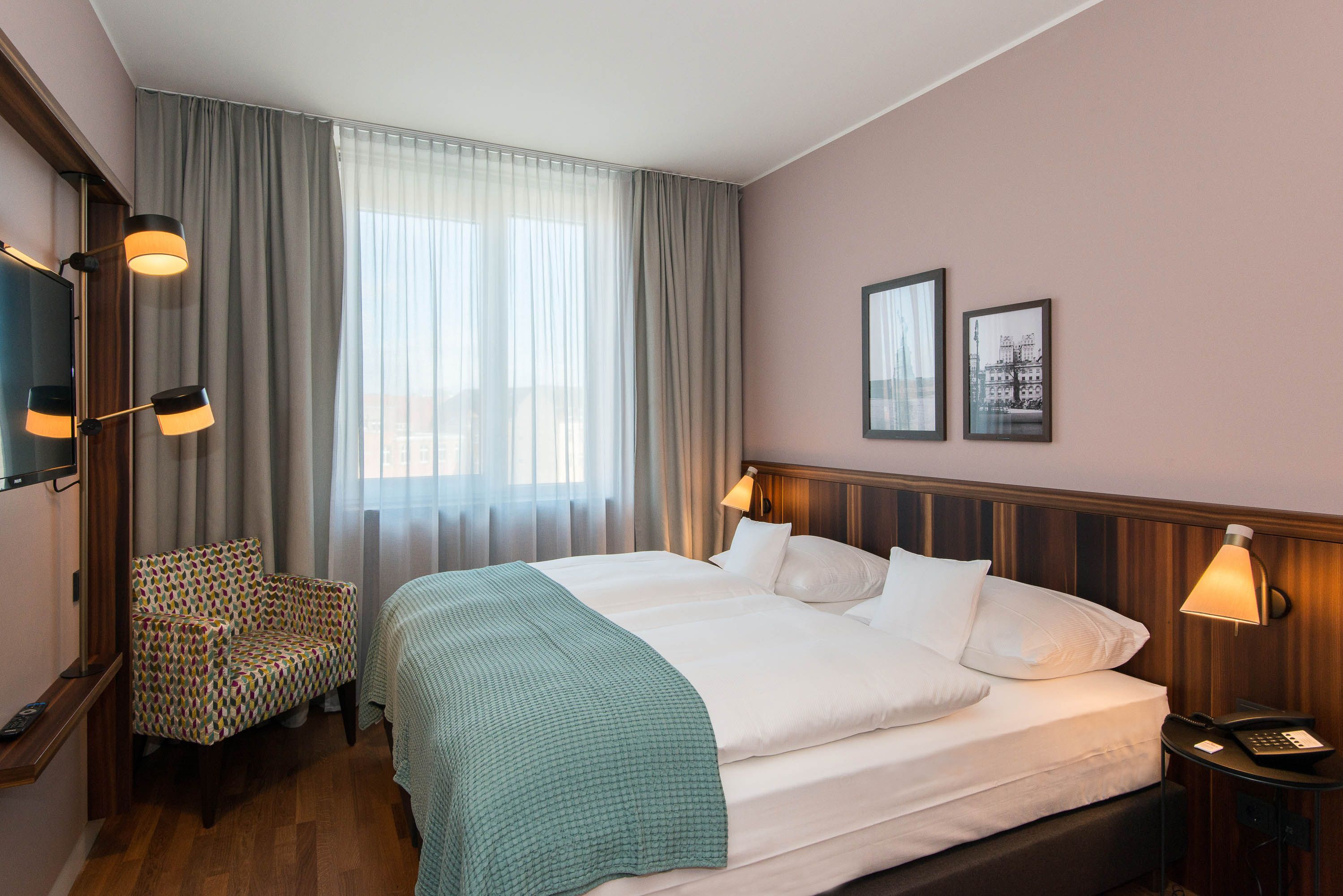 A hotel room with a large bed, a chair, a TV, and a bedside table with a lamp.