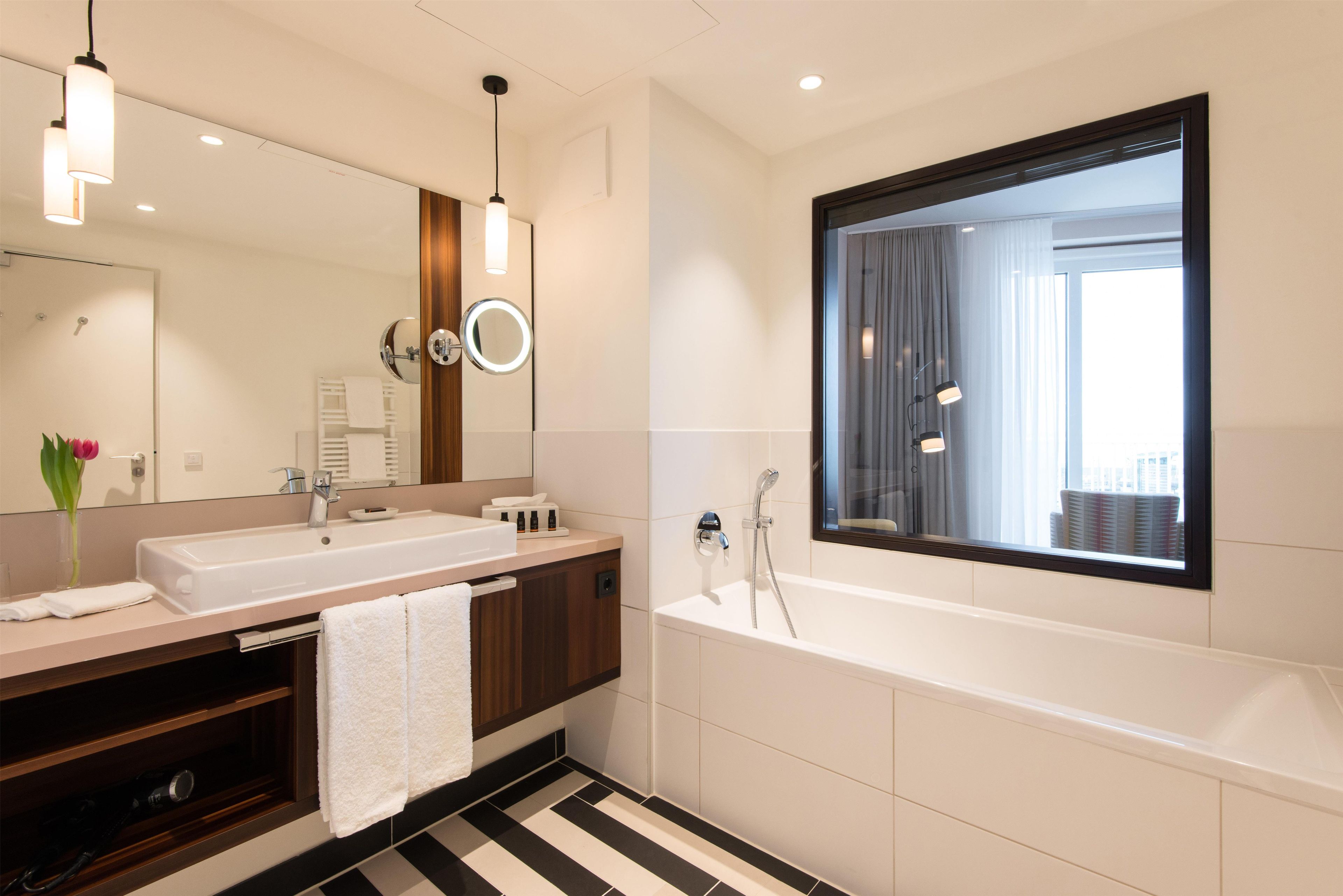 A modern bathroom with a large mirror, two sinks, a bathtub, and a window.