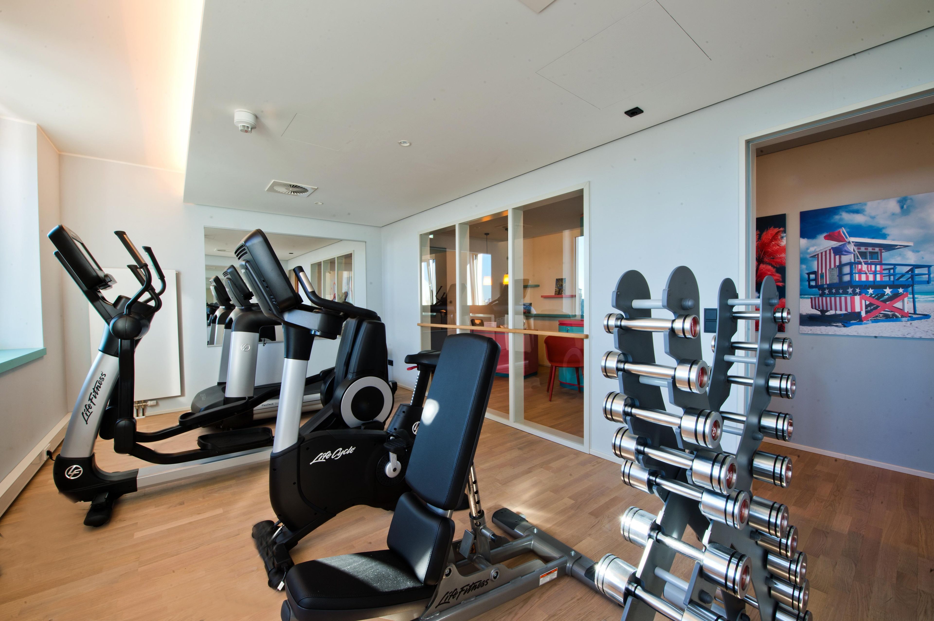 A gym with cardio machines, weightlifting equipment, and a dining area visible through glass doors.