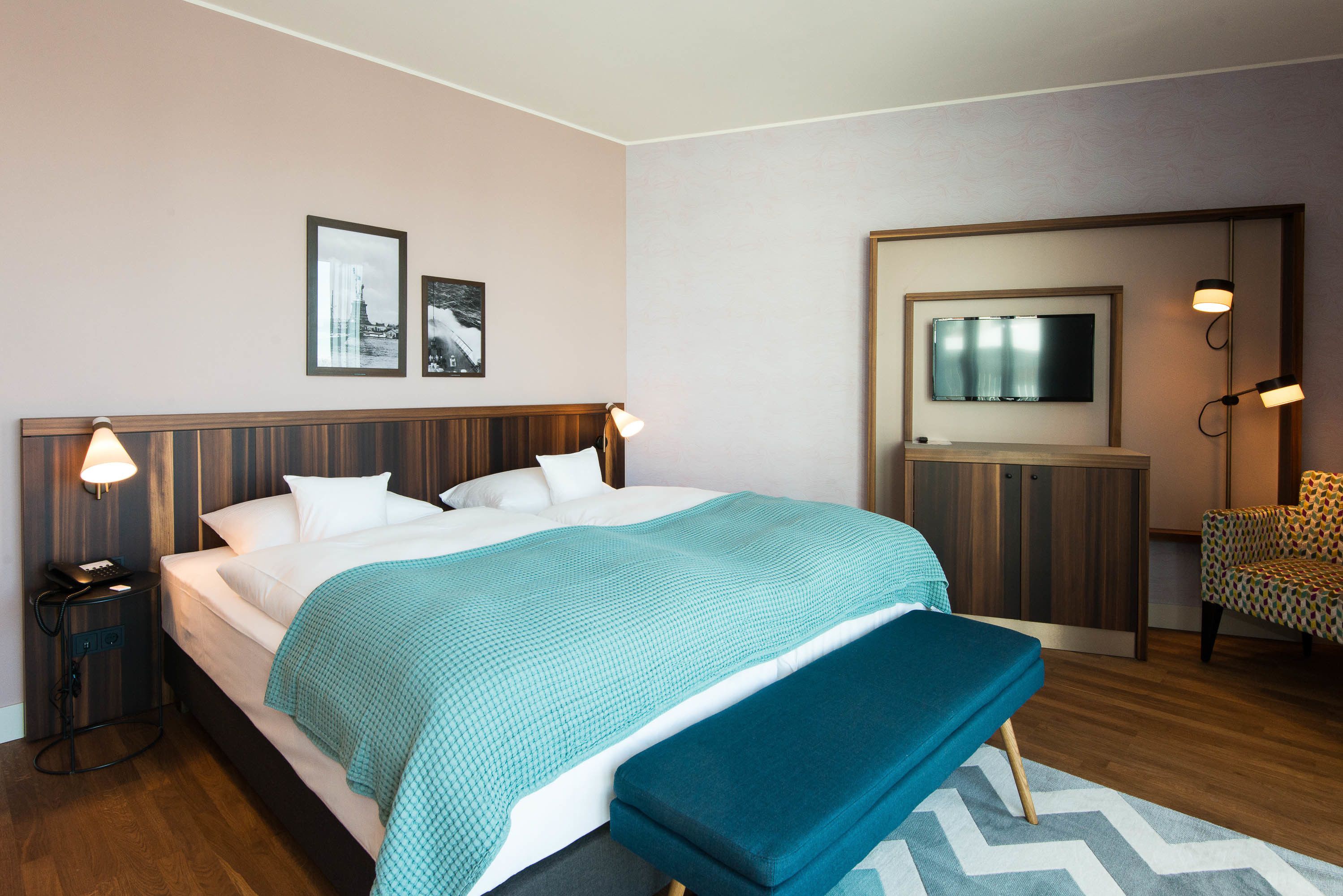 Quarto de hotel com cama, um banco, escrivaninha, televisão e cadeira.