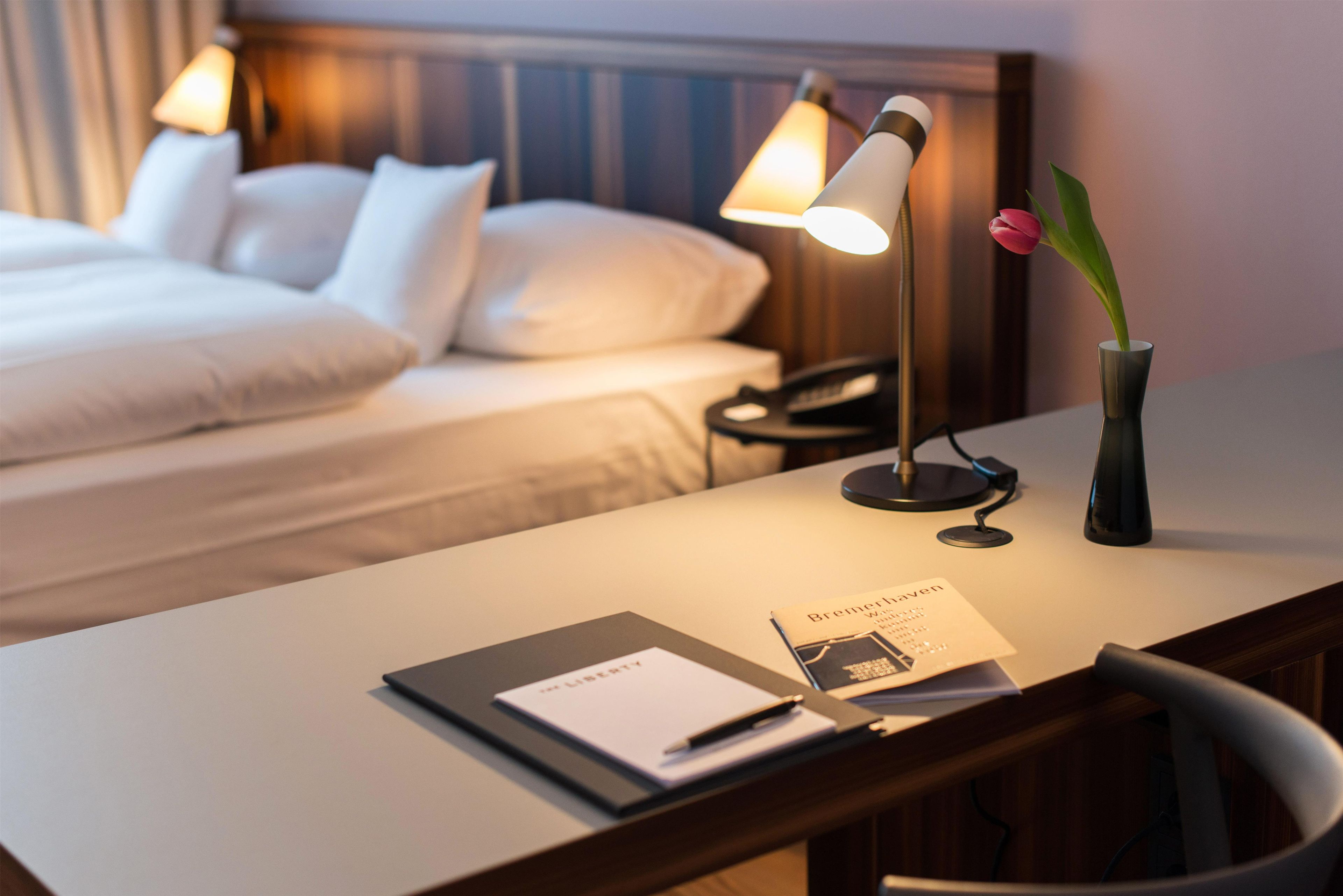 Habitación de hotel con una cama bien hecha, un escritorio con lámpara, un teléfono, un jarrón con una flor y un folleto sobre el escritorio.