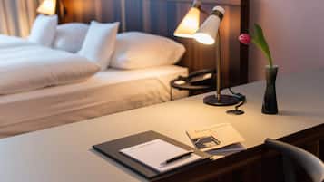 Habitación de hotel con una cama bien hecha, un escritorio con lámpara, un teléfono, un jarrón con una flor y un folleto sobre el escritorio.